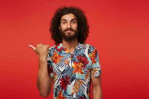 Portrait of pretty bearded brunette curly man smiling slightly to camera and showing aside with raised thumb, keeping lips folded while standing against red background, dressed in flowered shirt photo