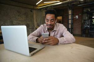 Positive handsome bearded man with dark skin wearing beige shirt, working remotely from coworking space, keeping mobile phone in hands and checking social networks photo