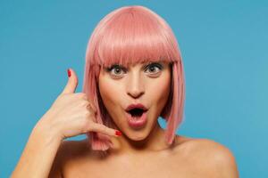 Close-up of positive green-eyed young pretty lady with trendy pink haircut rounding her mouth while looking cheerfully at camara, folding raised hand in handset, isolated over blue background photo