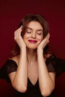 Pleased attractive young brown haired female lady with festive makeup wearing elegant black top with red dots while posing over claret background, smiling nicely with closed eyes photo