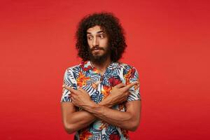 Puzzled young brunette curly bearded man in multi-colored shirt with floral print wrinkling forehead and looking confusedly aside, crossing hands with forefingers showing in different sides photo