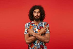 Severe handsome brunette male with lush beard showing in different directions with forefingers, frowning his eyebrows while looking at camera, isolated against red background photo