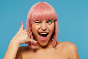 Indoor shot of joyful pretty young female with pink short hair closing one eye while looking aside and keeping her mouth wide opened, raising hand in call me gesture over blue background photo