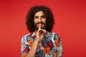 Cheerful pretty young brunette curly man with beard raising index finger in hush gesture, looking positively to camera with pleasant smile, isolated over red background in casual clothes photo