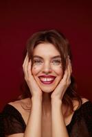 Charming blue-eyed young lady with brown wavy hair wearing silver stars on face and holding palms on her cheeks, smiling cheerfully to camera while posing against claret background photo