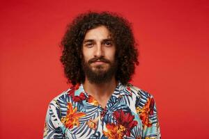 Close-up of attractive young brunette male with lush beard and curly hair looking positively to camera and smiling slightly, keeping lips folded while posing against red background photo