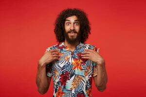 Joyful pretty brown-eyed brunette guy with long curly hair looking surprisedly to camera and keeping palms on his chest, posing against red background in shirt with floral print photo