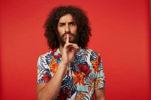 Serious brunette curly bearded guy in multi-colored flowered shirt keeping forefinger on his lips and frowing eyebrows, asking to keep silence, standing against red background photo