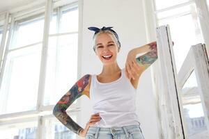 Happy young beautiful tattooed blonde lady with casual hairstyle leaning on wooden window while looking happily at camera, standing over bright studio on sunny day photo