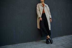 recortado foto de joven hipster mujer vestido en negro culottes, astuto suéter y beige zanja, posando terminado negro urbano pared en de moda ropa y cuero brogues