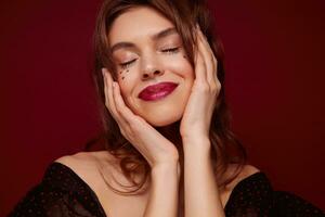 Enjoyable young brown haired pretty woman with red lips and little silver stars on her face smiling pleasantly with closed eyes, holding face with raised hands while posing against claret background photo