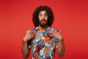 Amazed young brunette curly bearded man showing on himself with raised palms and looking to camera with wide eyes opened, dressed in multi-colored flowered shirt while standing over red background photo