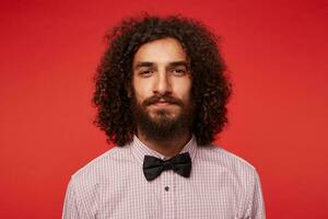 Close-up of young charming dark haired curly man with lush beard looking to camera with folded lips, wearing elegant clothes while standing against red background photo