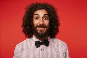 Surprised pretty young brunette male with beard rounding his brown eyes while looking to camera, raising eyebrows amazedly and wrinkling forehead while posing against red background photo