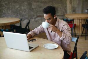 Young bearded dark skinned businessman with short haircut drinking coffee in city cafe while preparing materials on his laptop for meeting with clients, sitting at round wooden table in beige shirt photo