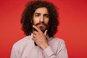 Thoughtful pretty dark haired curly guy with lush beard squinting pensively and holding his chin with raised hand, dressed in formal clothes while posing against red background photo