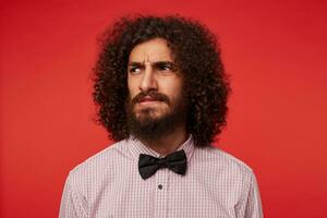 Studio shot of severe brunette curly man with beard looking aside and squinting with frowned eyebrows, dressed in elegant clothes while posing against red background photo