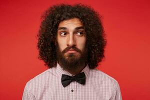 Confused young brunette curly male with beard wrinkling forehead while looking aside and twisting his mouth, standing over red background in elegant clothes photo