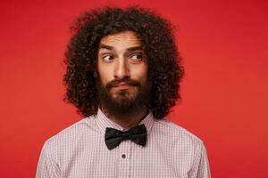 Young handsome bearded man with brown curly hair in checkered shirt and black bow-tie posing over red background, looking aside with doubting face and keeping lips folded photo