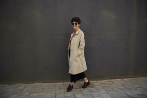 Outdoor shot of happy young pretty female with short haircut walking along black urban wall in stylish clothes and sunglasses, looking at camera cheerfully and smiling broadly photo