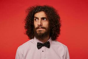 Portrait of young curly dark haired bearded male raising eyebrows and keeping lips folded while looking seriously aside, dressed in formal clothes while standing against red background photo