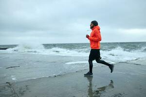 Outdoor photo of young active bearded man working out and running by seaside on stormy gray day, keeping hands at chest level and looking in front of himself