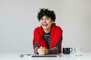Joyful young attractive curly brunette female dressed in striped pullover and red knitted sweater laughing happily while posing over white background with hands on countertop photo