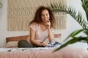 joven africano americano mujer con Rizado cabello, se sienta en el cama y toques mejilla, sonrisas y lee un nuevo revista, disfrutar gratis hora a hogar. foto