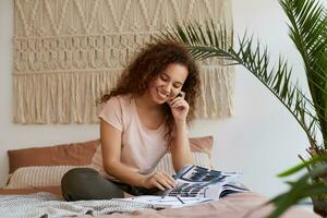 retrato de joven oscuro desollado dama con Rizado cabello, se sienta en el cama y toques mejilla, sonrisas y lee un nuevo revista, disfrutar gratis hora a hogar. foto