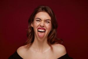 Close-up of joyful excited young brunette woman with silver stars on her face showing tongue to camera and squinting her eyes, posing over claret background in top with opened shoulders photo