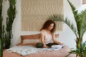 Young positive dark skinned woman with curly hair, sits on the bed and reads a favorite magazine, enjoy sunny morning at home and free time. photo