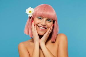 retrato de joven positivo bonito mujer vistiendo blanco flor en su corto rosado pelo mientras posando terminado azul fondo, participación palmas en su las mejillas y sonriente en general a cámara foto