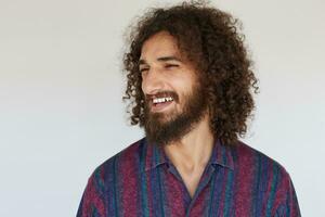 Cheerful handsome bearded young man with dark curly hair looking aside and laughing happily, being in high spirit while standing over white background in casual clothes photo