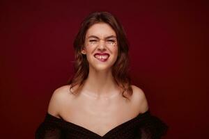 Portrait of young lovely positive female with brown hair frowning her face and biting underlip while looking at camera, dressed in elegant black top with red dots over claret background photo