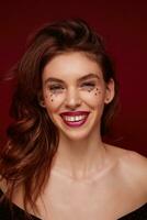 Close-up of young pretty brunette lady with evening makeup looking happily at camera and smiling widely, being in high spirit while posing against claret background photo