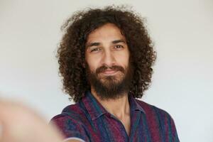 atractivo positivo morena Rizado hombre con barba participación cámara y sonriente atentamente mientras haciendo autofoto, vistiendo a rayas multicolor camisa mientras posando terminado blanco antecedentes foto