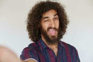 Cheerful lovely dark haired curly guy with lush beard winking to camera with wide mouth opened, wrinkling forehead while posing against white background in casual clothes photo