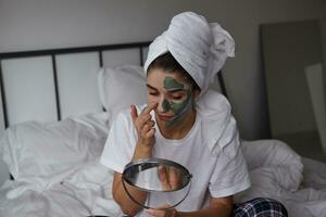 Indoor photo of young dark haired lady with white towel on her head holding mirror in her hand and applying mask on her face, dressed in casaul clothes while posing over bedroom interior