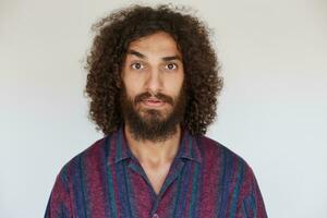 Puzzled curly brown-eyed brunette male with beard looking confusedly to camera and raising eyebrow, keeping hands down while standing over white background photo