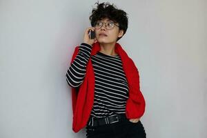 Attractive young stylish dark haired curly female in eyewear leaning on white wall while making call with her smartphone, dressed in striped pullover, red woolen sweater and black trousers photo