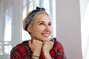 de cerca de joven contento rubia en venda propensión su cara en elevado manos y sonriente alegremente, siendo en bonito estado animico mientras en pie en frente de grande ventana foto