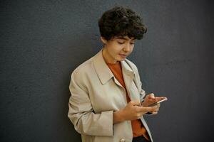 Attractive young curly brunette lady with short haircut standing over city environment in trendy outfit, holding mobile phone in her hands and looking at screen with folded lips photo