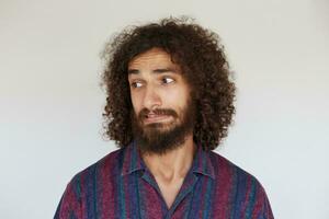 Studio shot of handsome young brunette curly man with beard looking aside with confused face and twisting his mouth, dressed in striped multi-colored shirt against white background photo