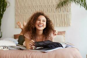 Portrait of young laughing dark skinned woman with curly hair, lies on the bed and reads a magazine, enjoy sunny free day, looks away and broadly smiles. photo