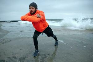 Young dark haired handsome bearded model with headphones keeping hands in front of himself and frowning eyebrows while looking ahead, doing morning workout over seaside photo