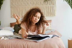 Young thinking african american woman with curly hair, lies on the bed, reads an article in a magazine in a concentrated manner. photo