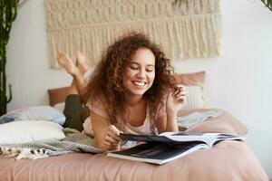 Portrait of young positive african american lady with curly hair, lies on the bed and reads a new issue of magazine, enjoy free day, broadly smiles and looks happy. photo