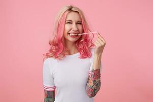 foto de joven sonriente hermosa rosado peludo dama con tatuado manos, usa en blanco camiseta, disfruta fresa burbuja chicle, mira a el cámara soportes terminado rosado antecedentes.