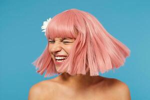 Pleasant looking young lovely lady with short pink haircut waving her head while posing over blue background, laughing happily with closed eyes, having white flower in her hair photo