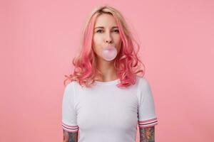 Portrait of young beautiful pink haired lady with tattooed hands, wears in white t-shirt, blows gum ball, looks at the camera stands over pink background. photo